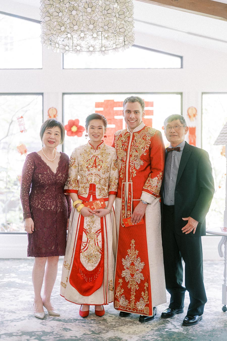 Miffy and Rawle in red Chinese wedding attire standing with two parents by bright windows.