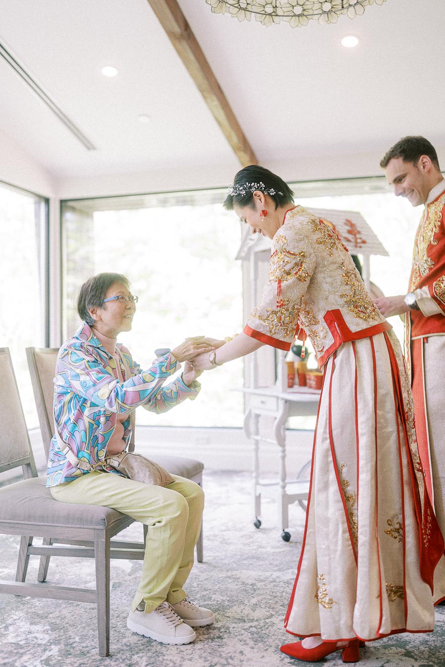 Bride in red and gold tea ceremony dress offers tea to a seated elder while groom stands beside her.