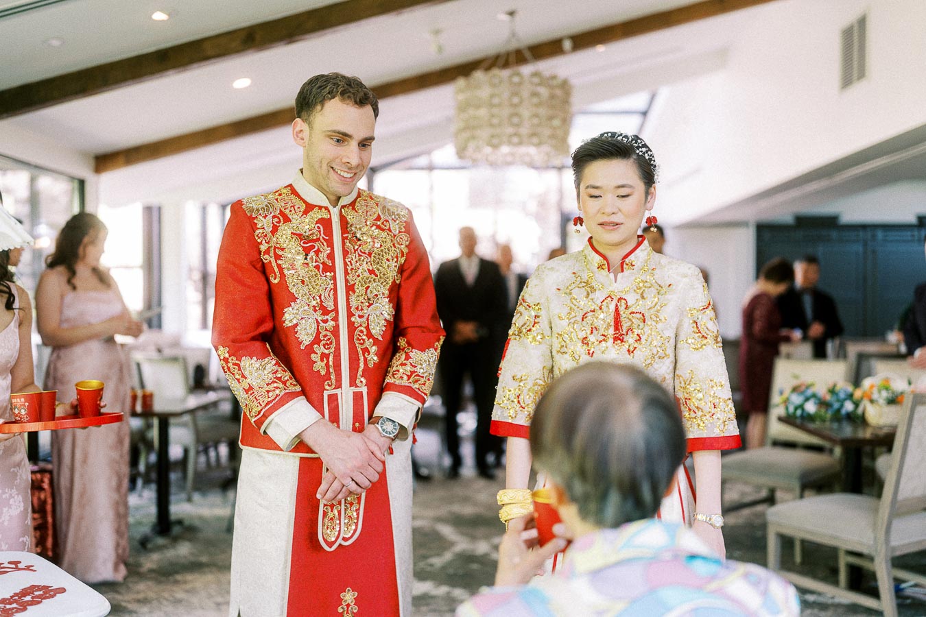 Miffy and Rawle stand in red and gold Chinese wedding attire before an elder holding a tea cup.