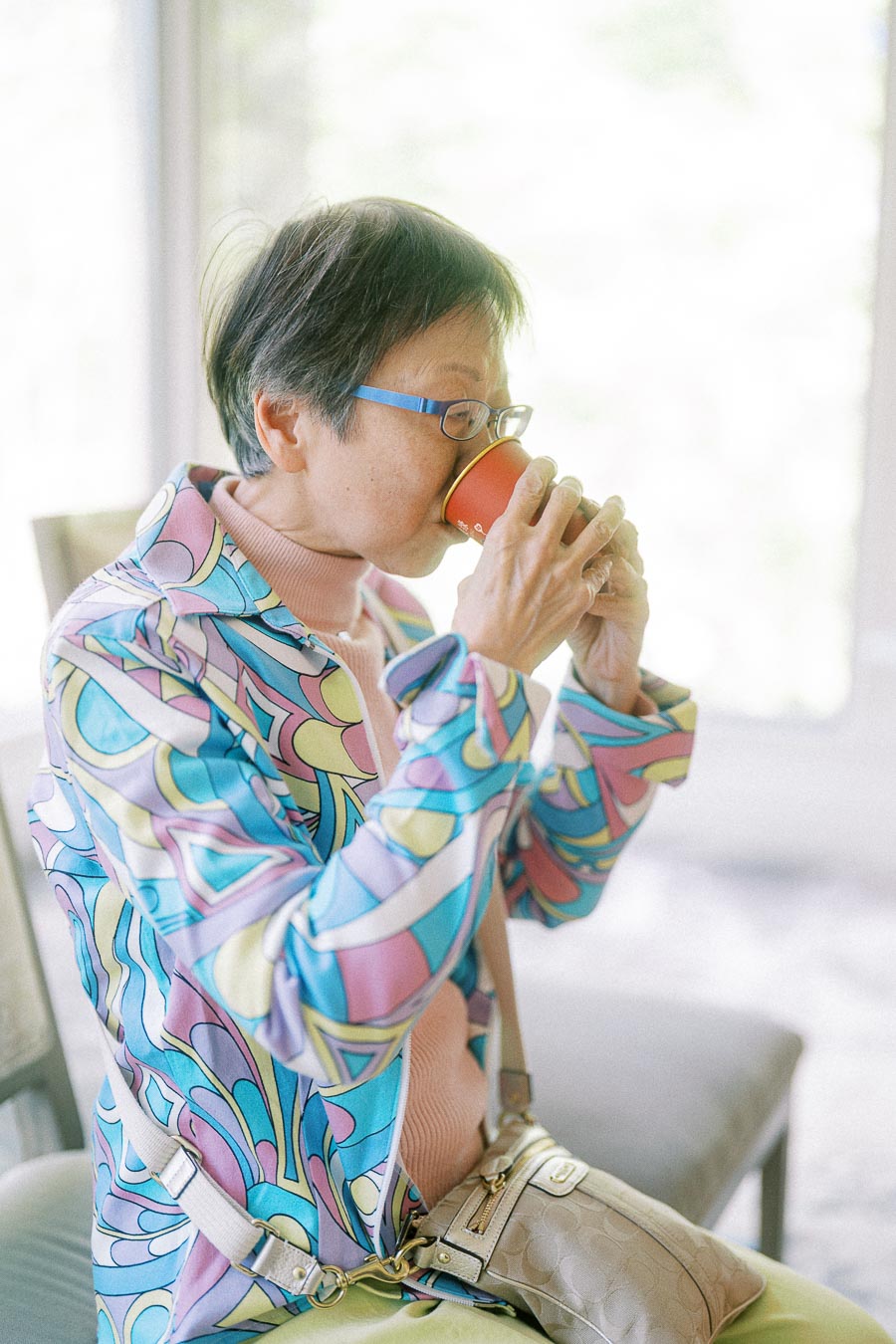 Older woman in glasses and a colorful patterned jacket drinks from a red cup beside a bright window.