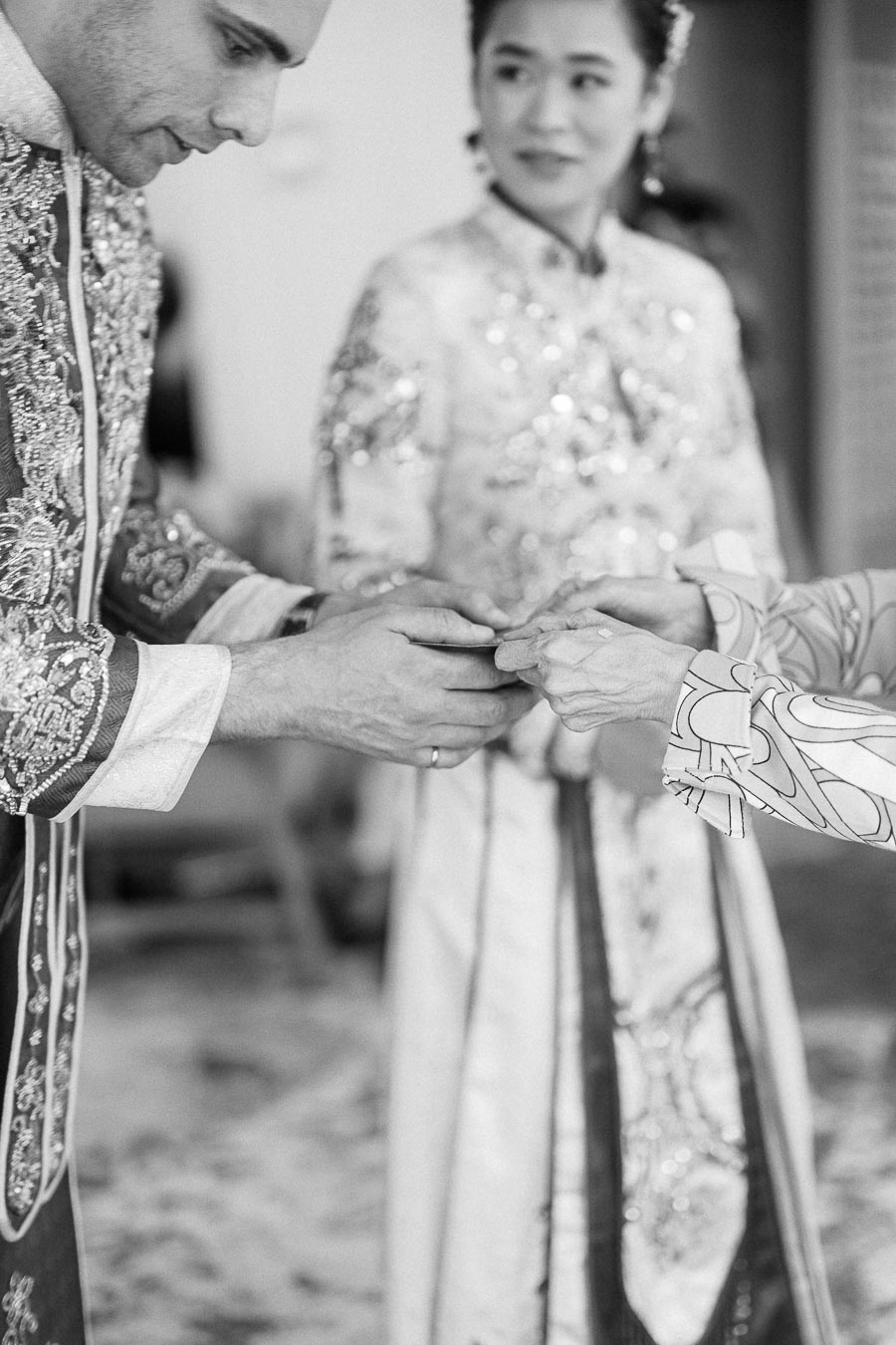 Groom in embroidered attire places a ring on an elder’s hand during a Chinese tea ceremony as the bride watches.