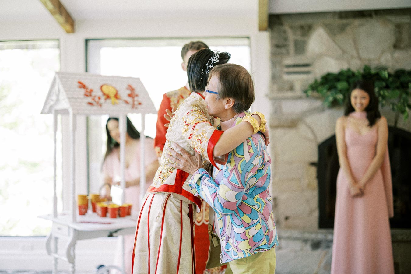 Bride in a Chinese wedding outfit hugs a guest during a tea ceremony inside a bright stone-walled room.