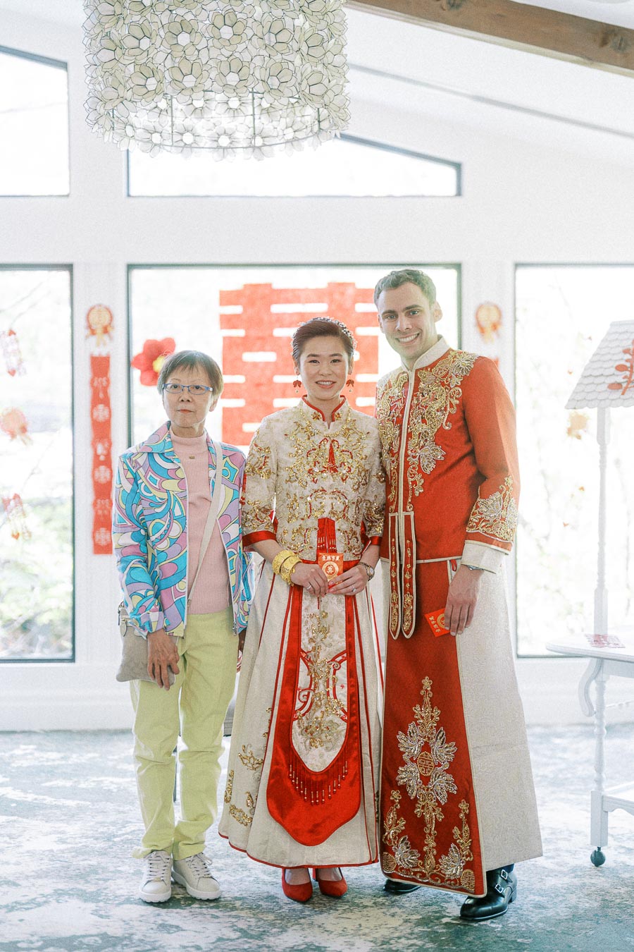 Miffy and Rawle in ornate Chinese wedding attire standing with a woman beside a red double happiness backdrop.