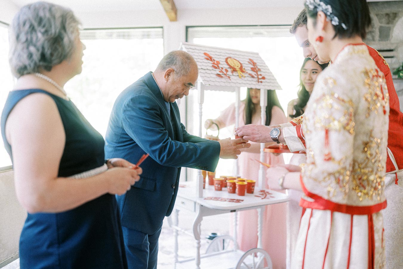 Father places a ring on the groom’s hand during a Chinese tea ceremony beside the bride in red and gold attire.