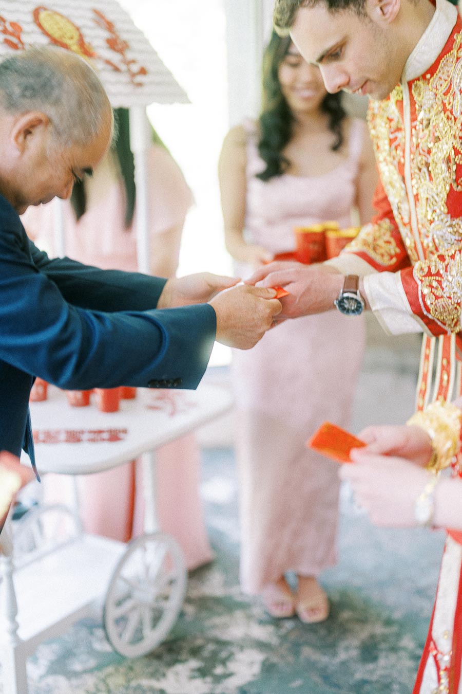 Elder places a red envelope in Rawle’s hand during a Chinese tea ceremony with bridesmaids nearby.