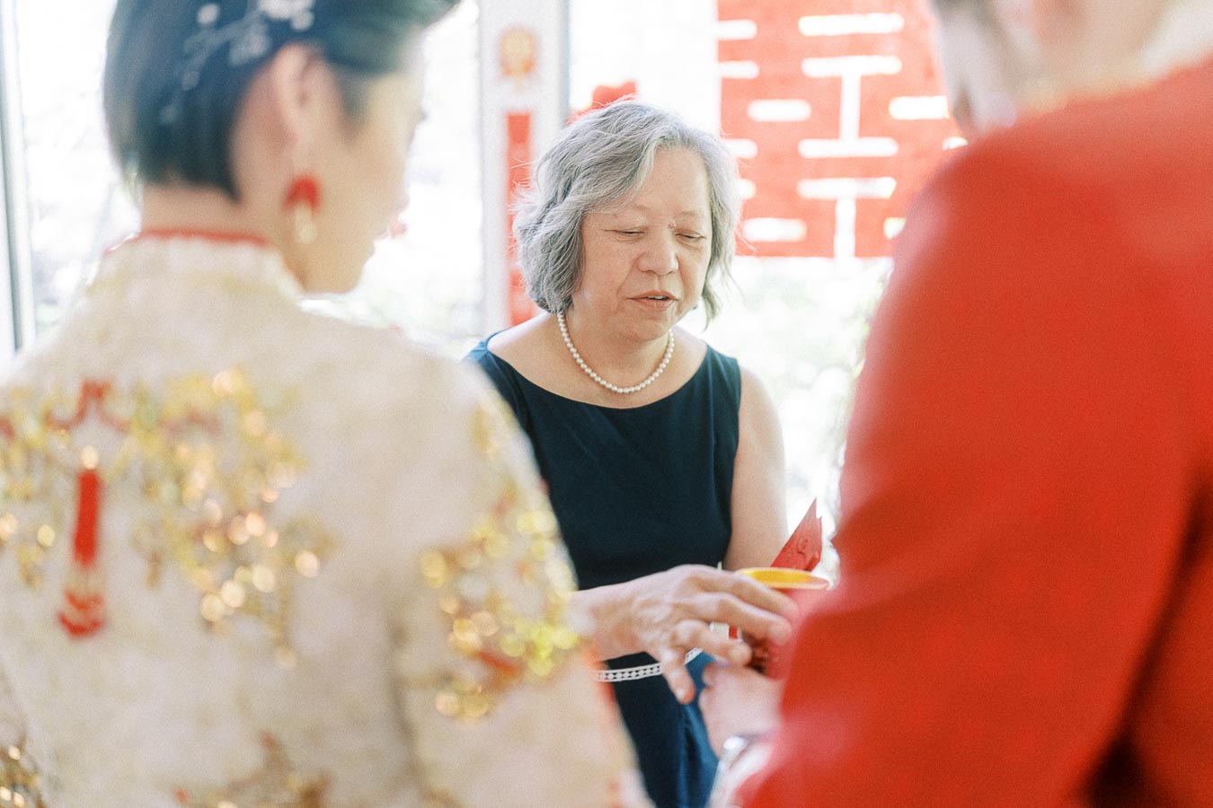 Elder woman in black offers a red envelope during a Chinese tea ceremony with the bride in embroidered attire.