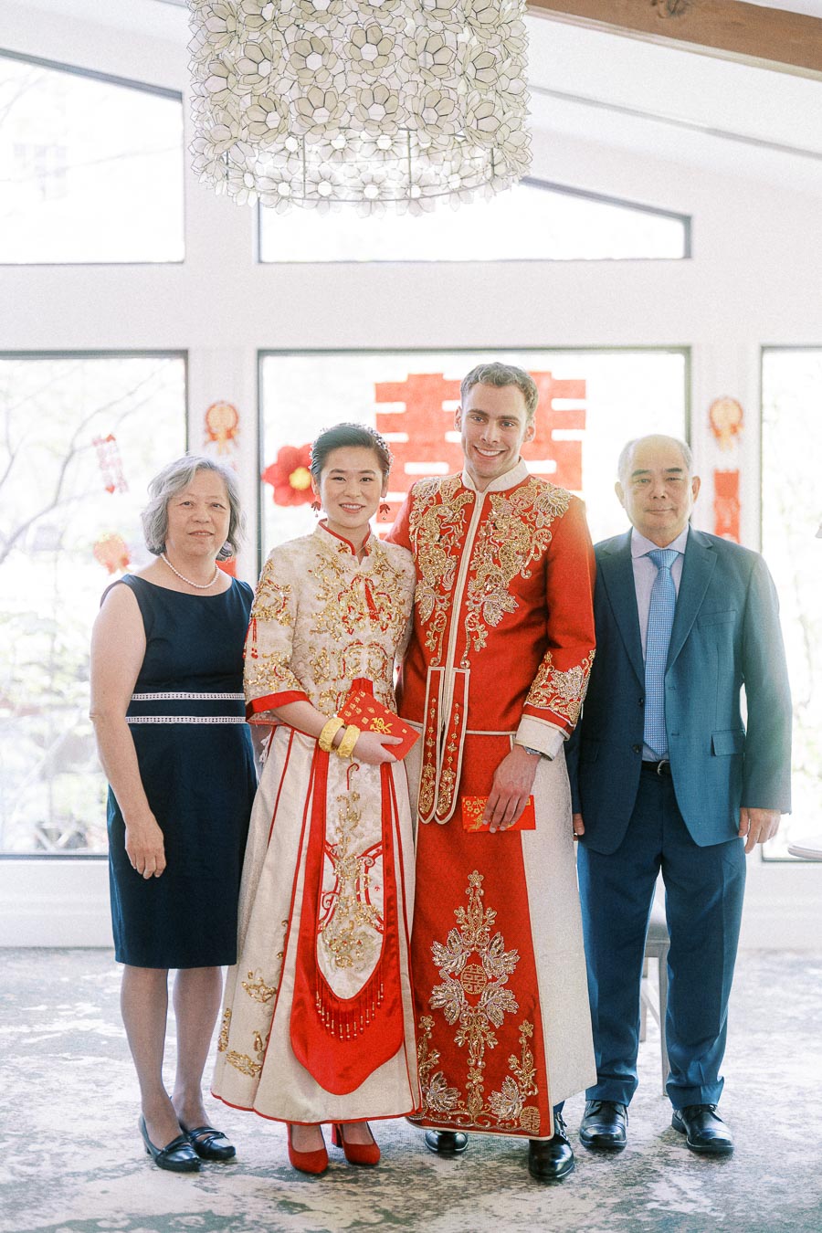 Miffy and Rawle stand with two family members in red and gold Chinese wedding attire indoors.