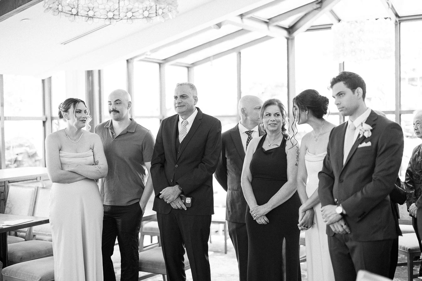 Wedding guests and couple stand together in a bright ceremony room with large windows and chandeliers.