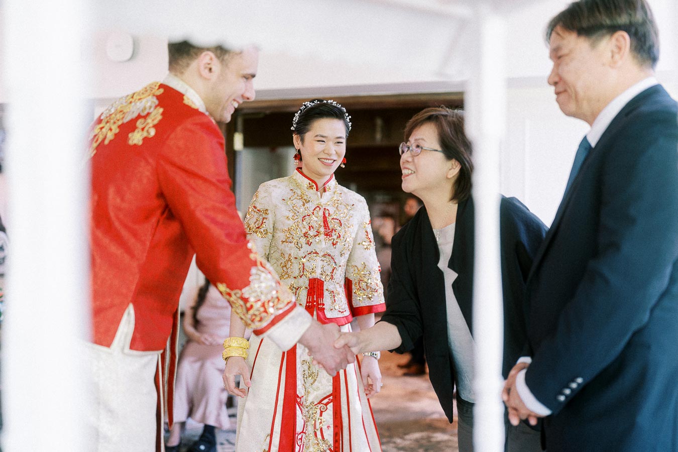 Groom in red traditional jacket shakes hands with an older woman as bride smiles beside them during a tea ceremony.