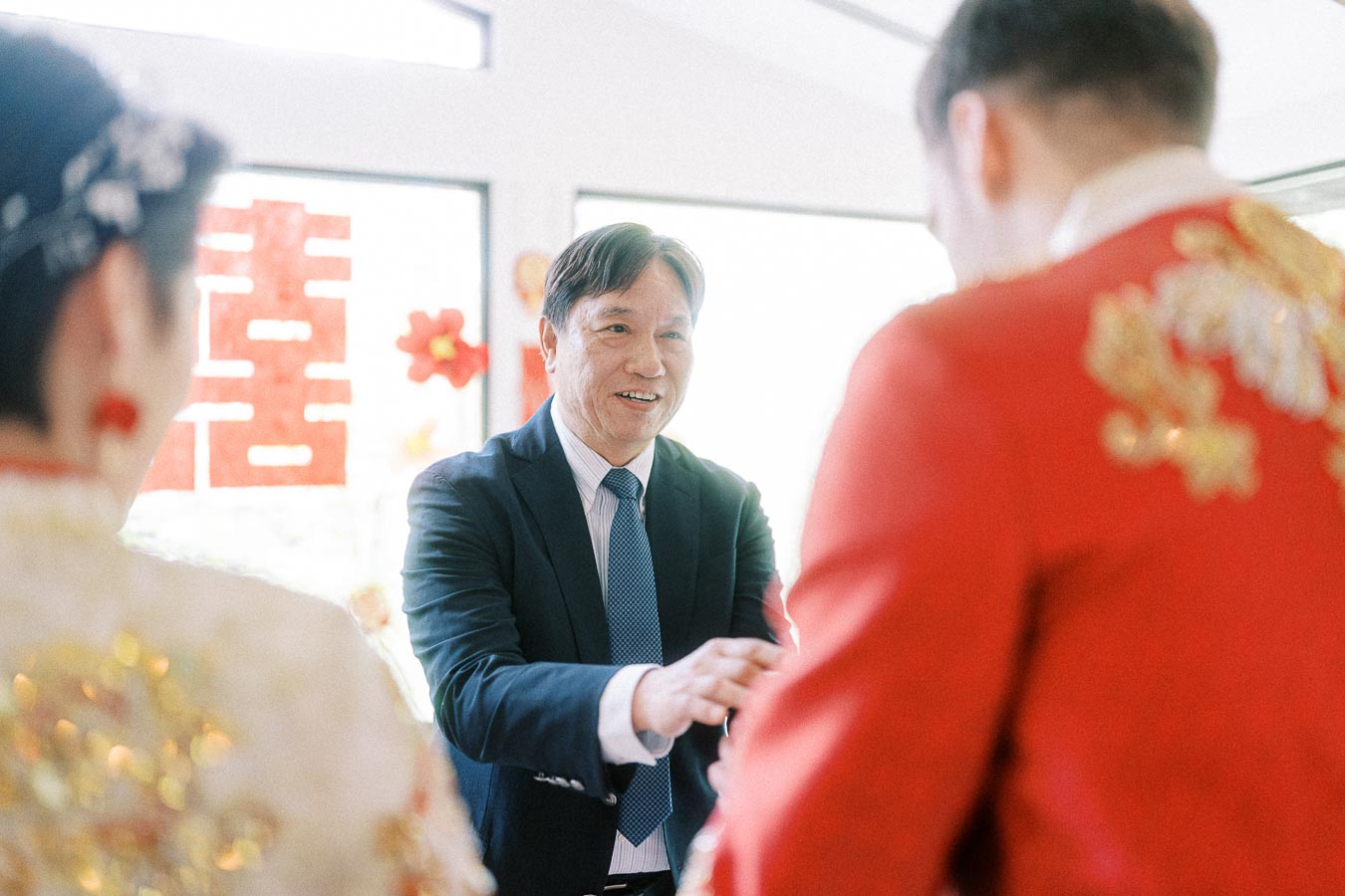 Father shakes hands with groom in red traditional attire during a Chinese tea ceremony.