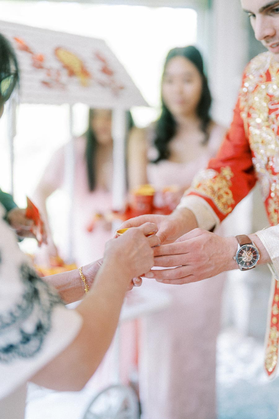 Hands exchange a teacup during a Chinese tea ceremony, with the groom in a red embroidered jacket.