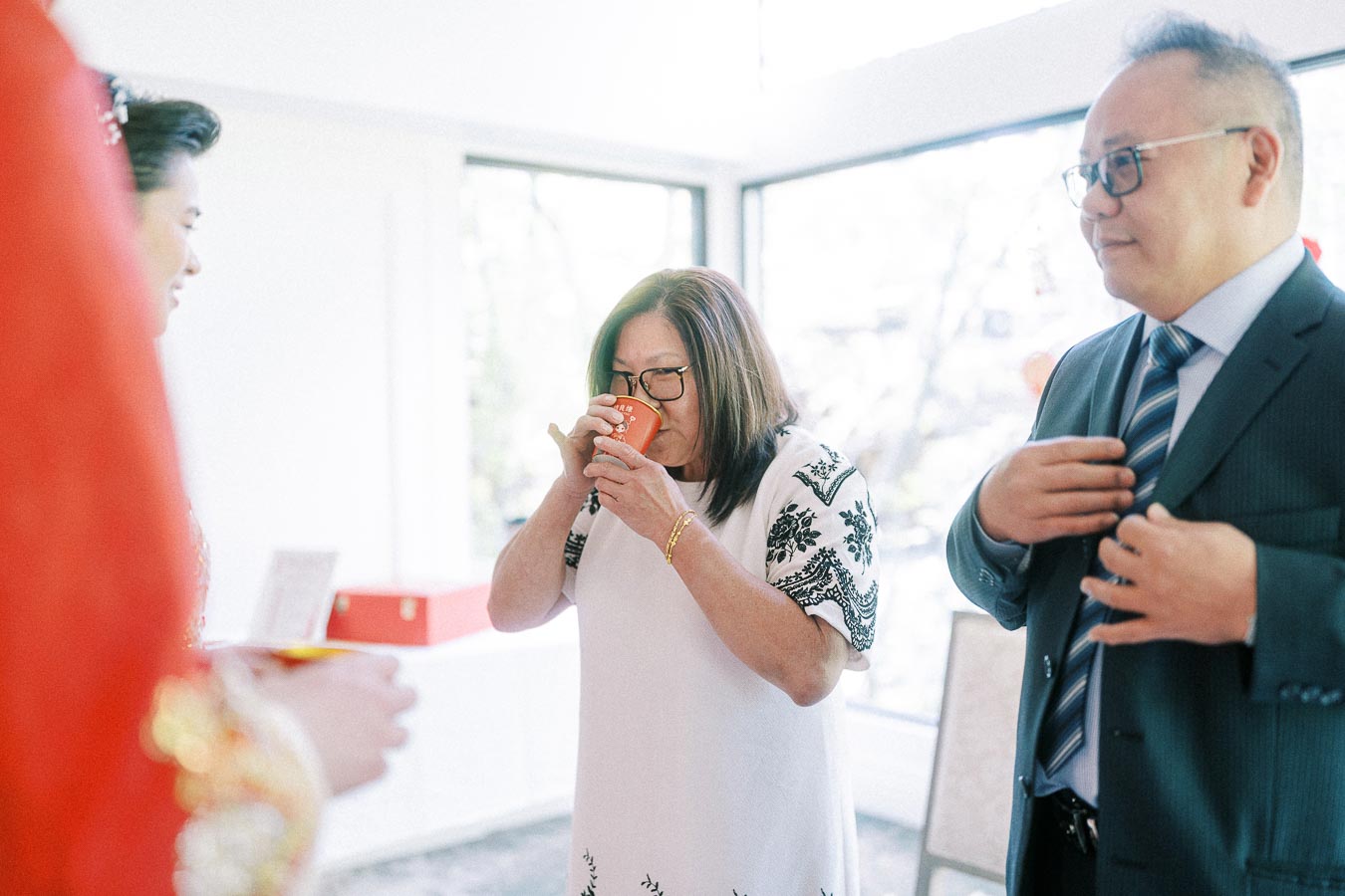 Woman drinks tea from a red cup as a suited man watches during a Chinese tea ceremony indoors.