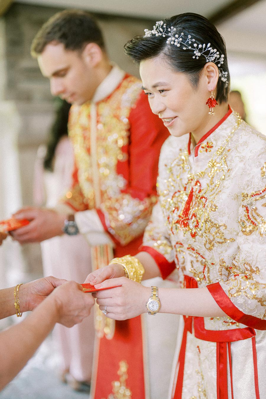 Miffy and Rawle in red and gold Chinese wedding attire offering a red envelope during tea ceremony