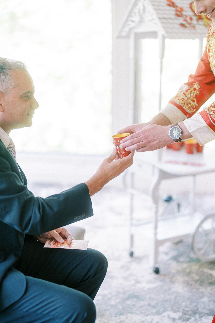 Groom in embroidered red jacket offers tea to a seated man during a Chinese tea ceremony.