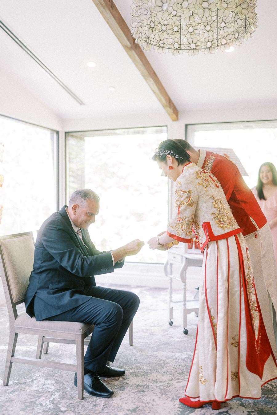 Bride and groom in red traditional wedding attire serve tea to a seated man during a Chinese tea ceremony.