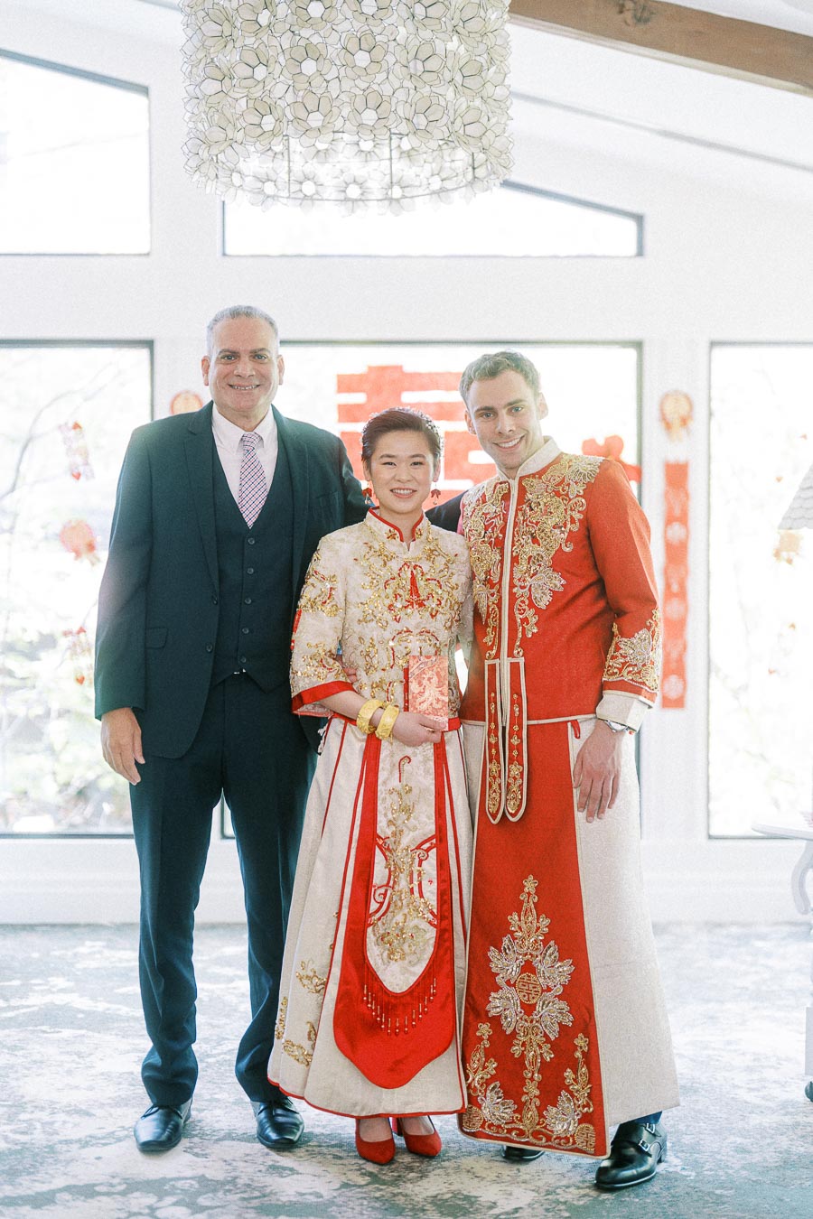 Miffy and Rawle pose with a suited man in ornate red and gold Chinese wedding attire indoors.