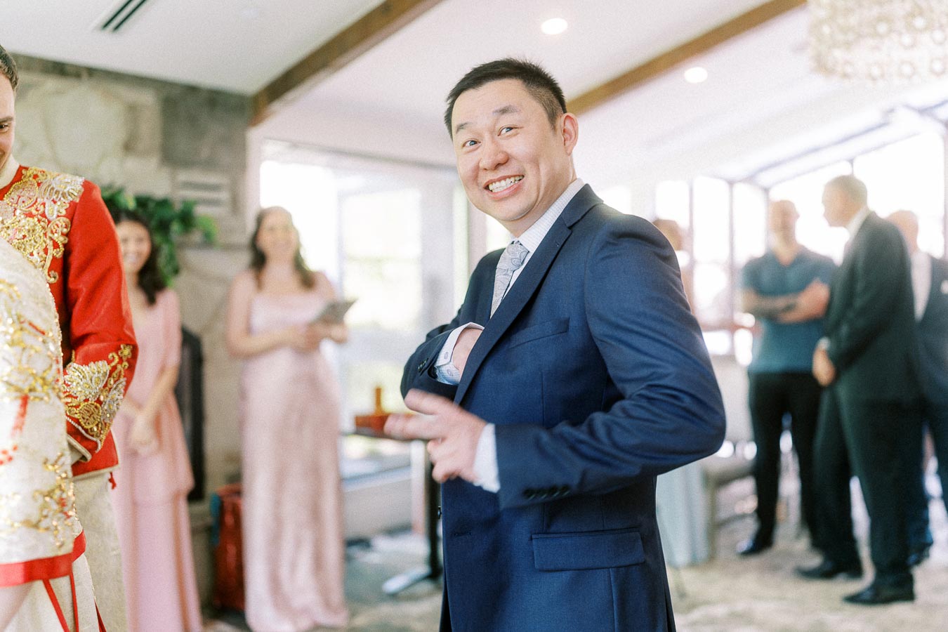 Groom in a navy suit smiles while adjusting his jacket, with wedding guests and a red ceremonial outfit nearby.