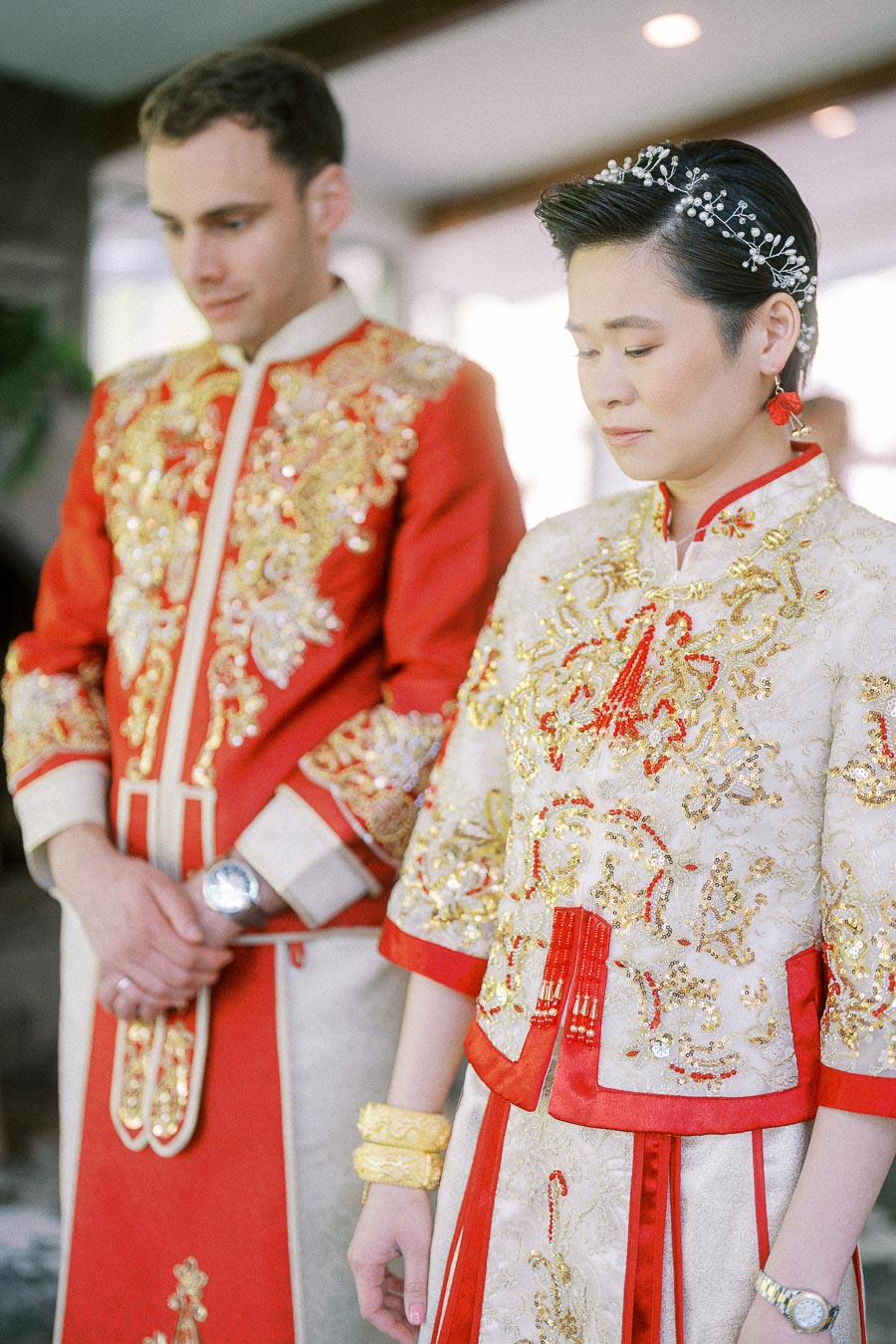 Miffy and Rawle stand in ornate red and gold Chinese wedding outfits with heads bowed during their tea ceremony.