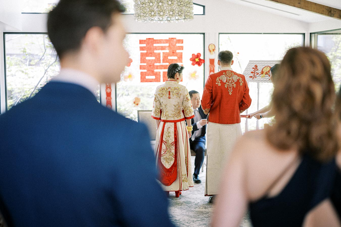 Couple in red and gold Chinese wedding attire stands before elders during a tea ceremony