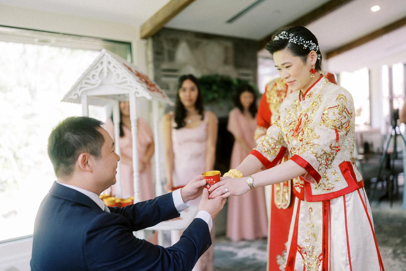 Groom kneels and offers a red teacup to bride in ornate red and gold Chinese wedding attire.