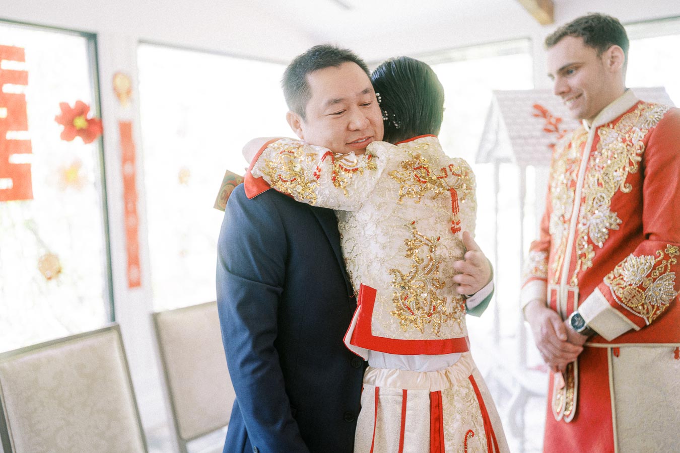 Groom in a navy suit hugs a person in ornate cream and red Chinese wedding attire indoors.