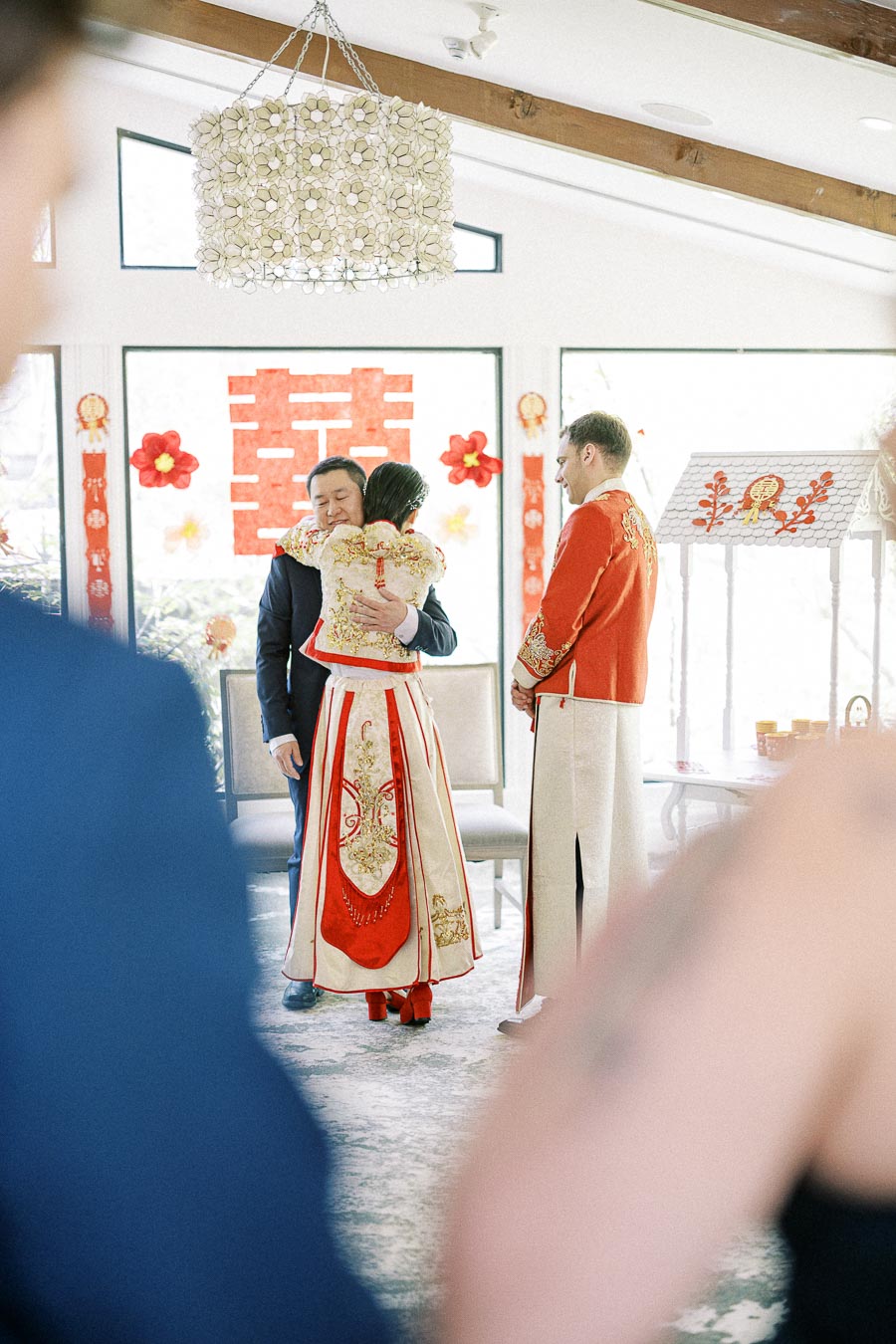 Bride in red and gold Chinese wedding dress hugs a man during a tea ceremony as the groom stands nearby.