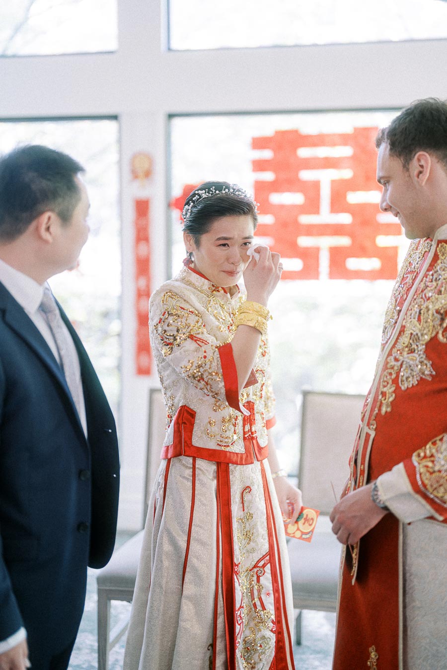Bride in embroidered Chinese wedding dress wipes a tear beside the groom and a guest under a double happiness sign.