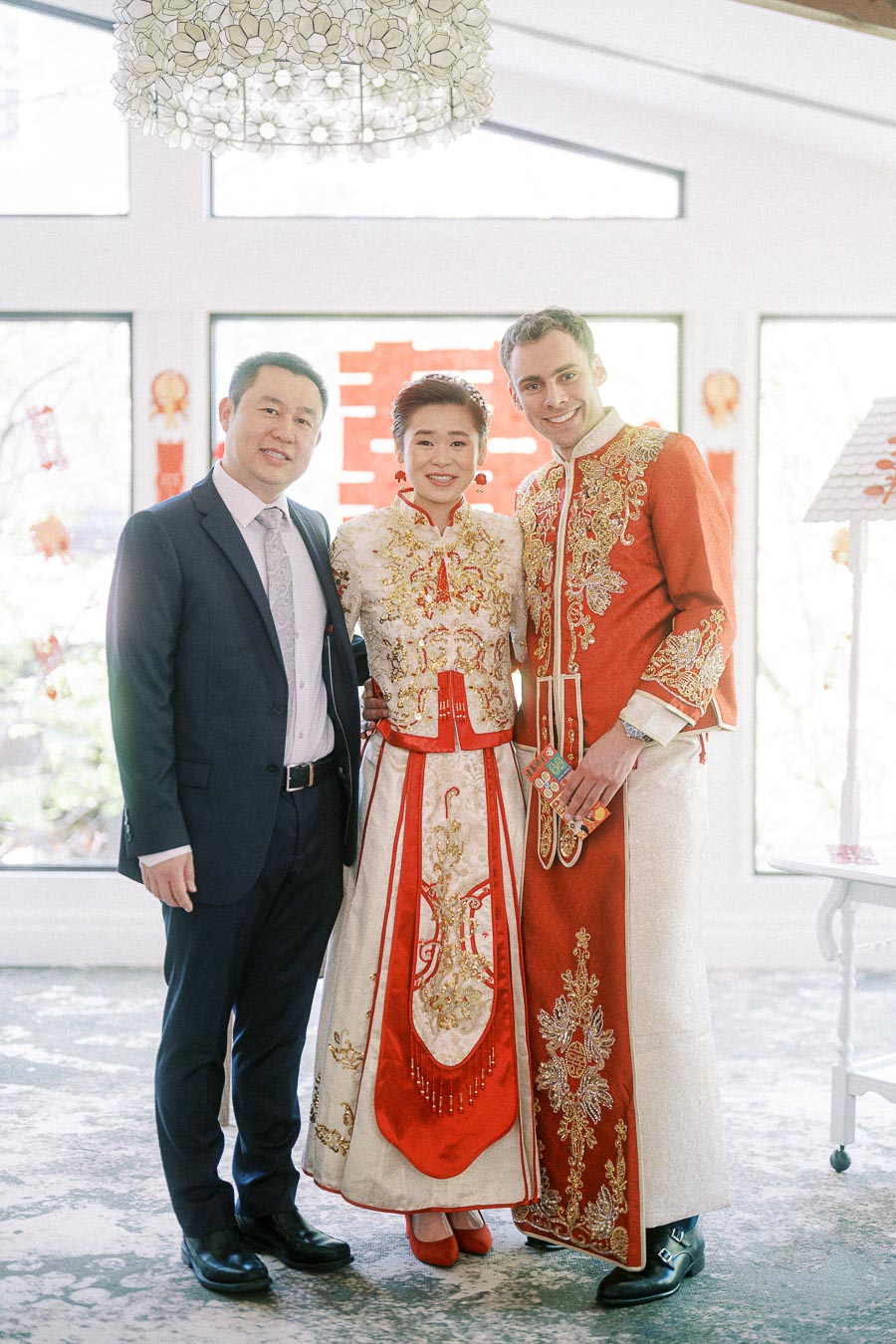 Miffy and Rawle in red Chinese wedding attire posing with a suited man beside bright windows