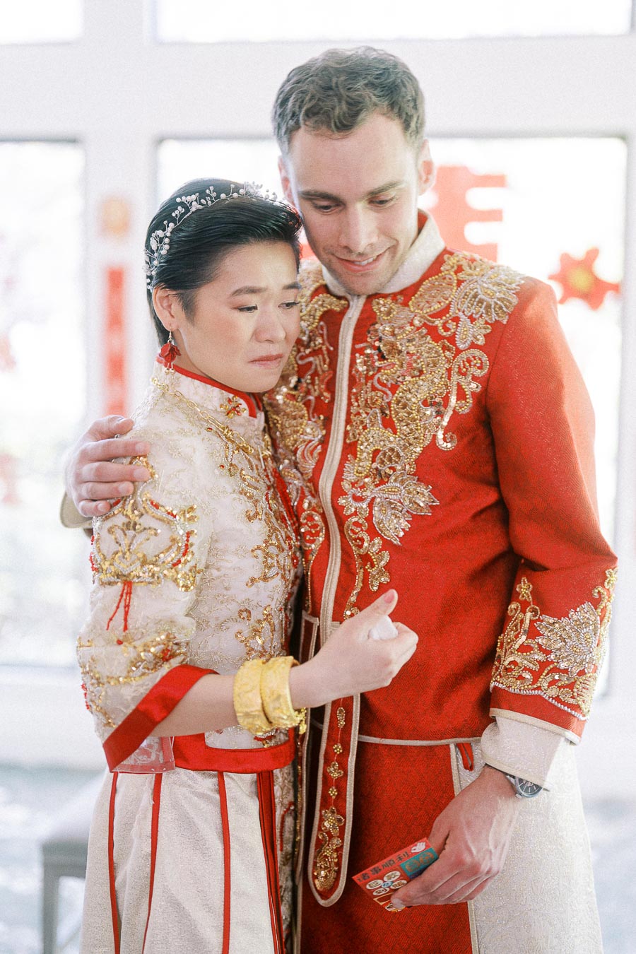Miffy and Rawle embrace in ornate red and gold Chinese wedding outfits during their Ancaster Mill celebration.
