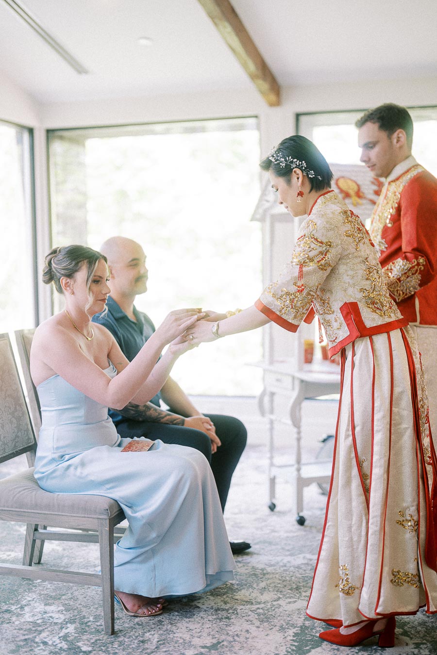 Bride in red and gold tea ceremony dress offers tea to seated woman while groom stands behind her.