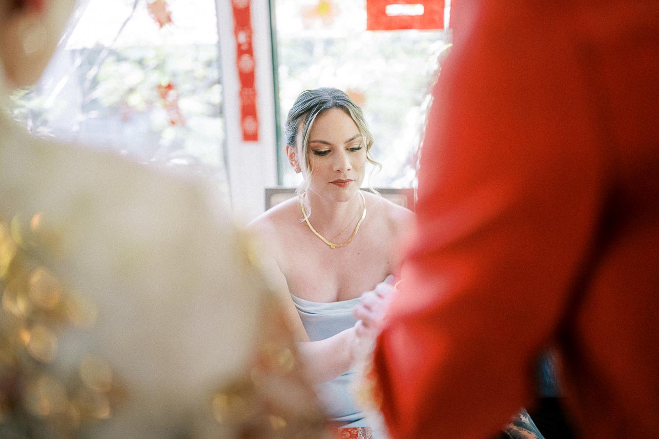 Bride Miffy sits indoors between red Chinese tea ceremony decorations, looking down with hands clasped.