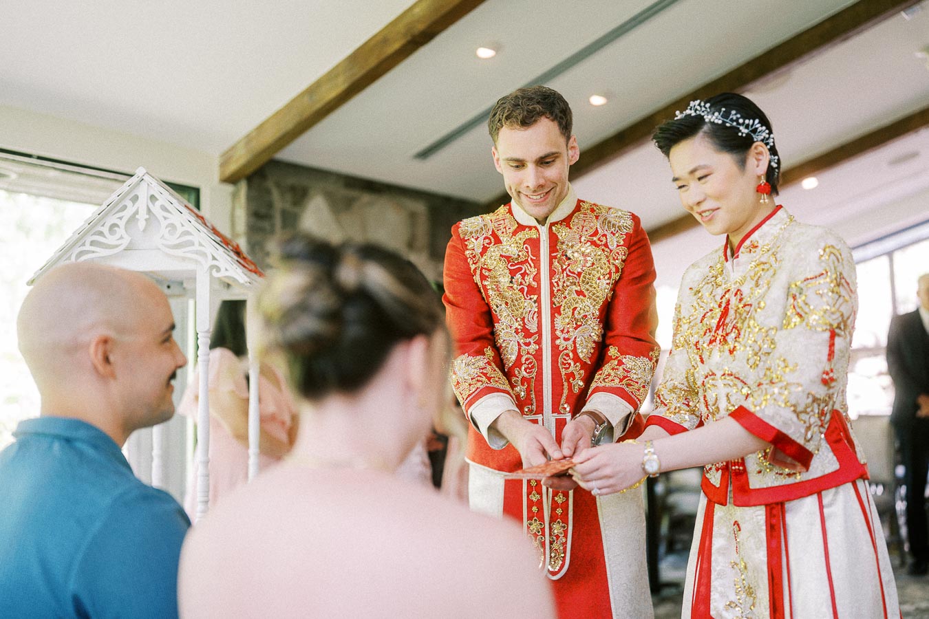 Miffy and Rawle in red embroidered Chinese wedding outfits serving tea to seated family members.