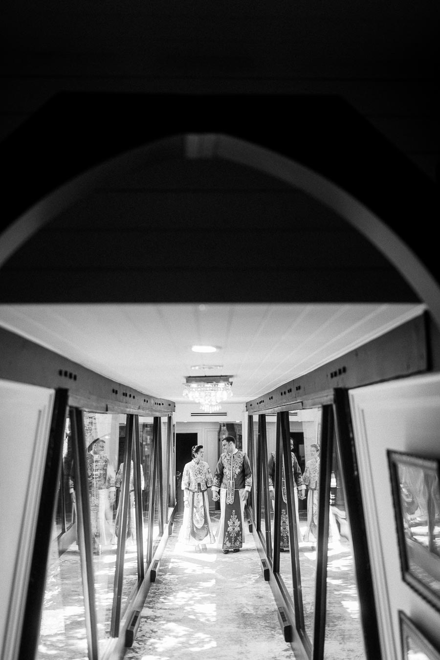 Miffy and Rawle walk hand in hand through a mirrored hallway in traditional wedding attire.