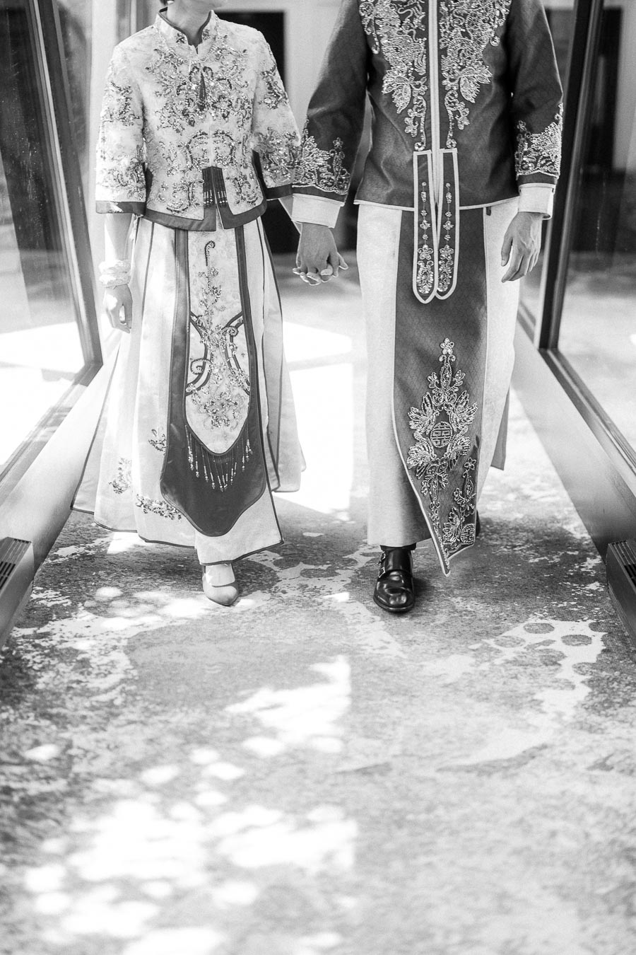 Couple walking hand in hand in embroidered Chinese wedding outfits in a bright corridor