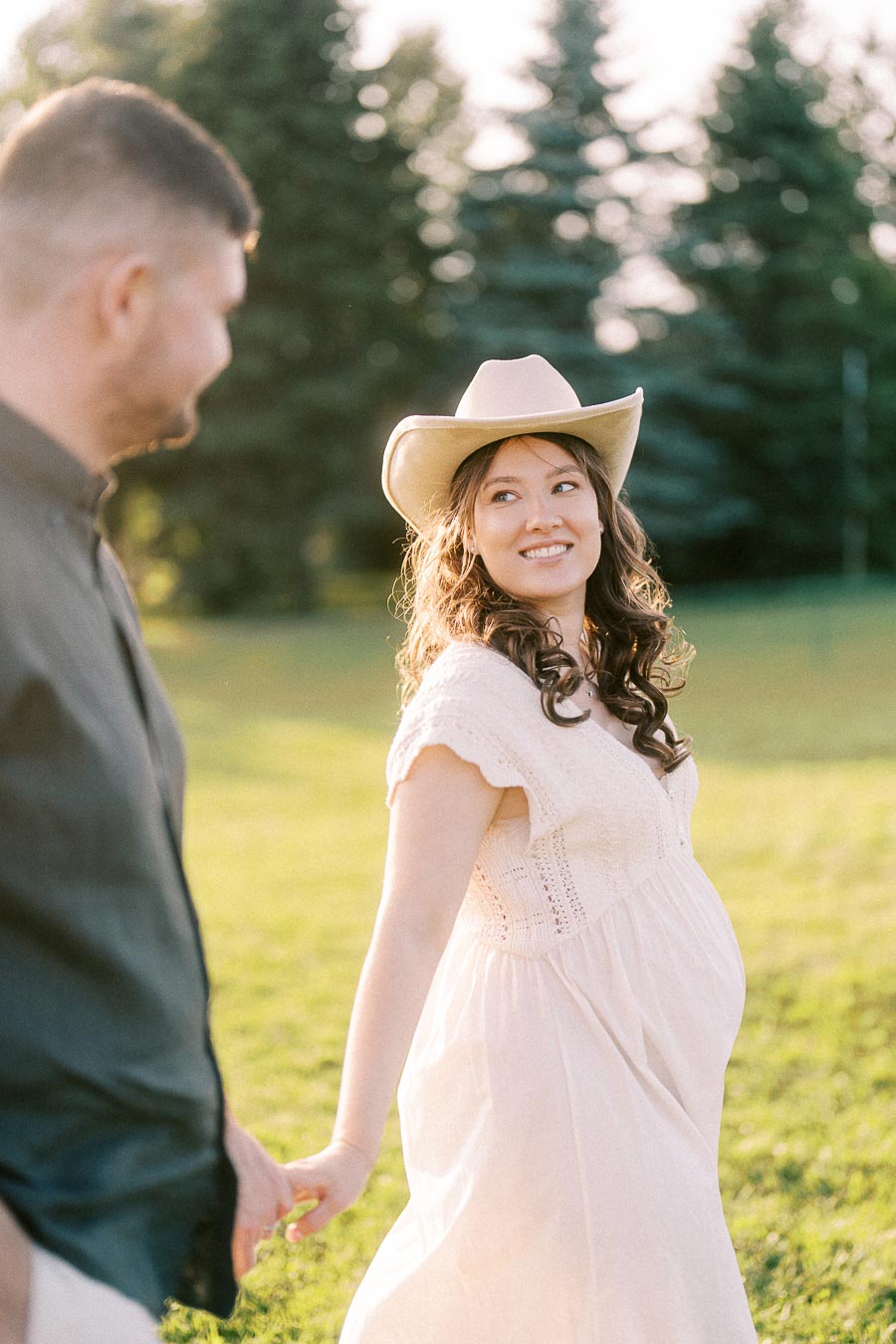 Pregnant woman in cowboy hat holding hands with partner in a grassy field