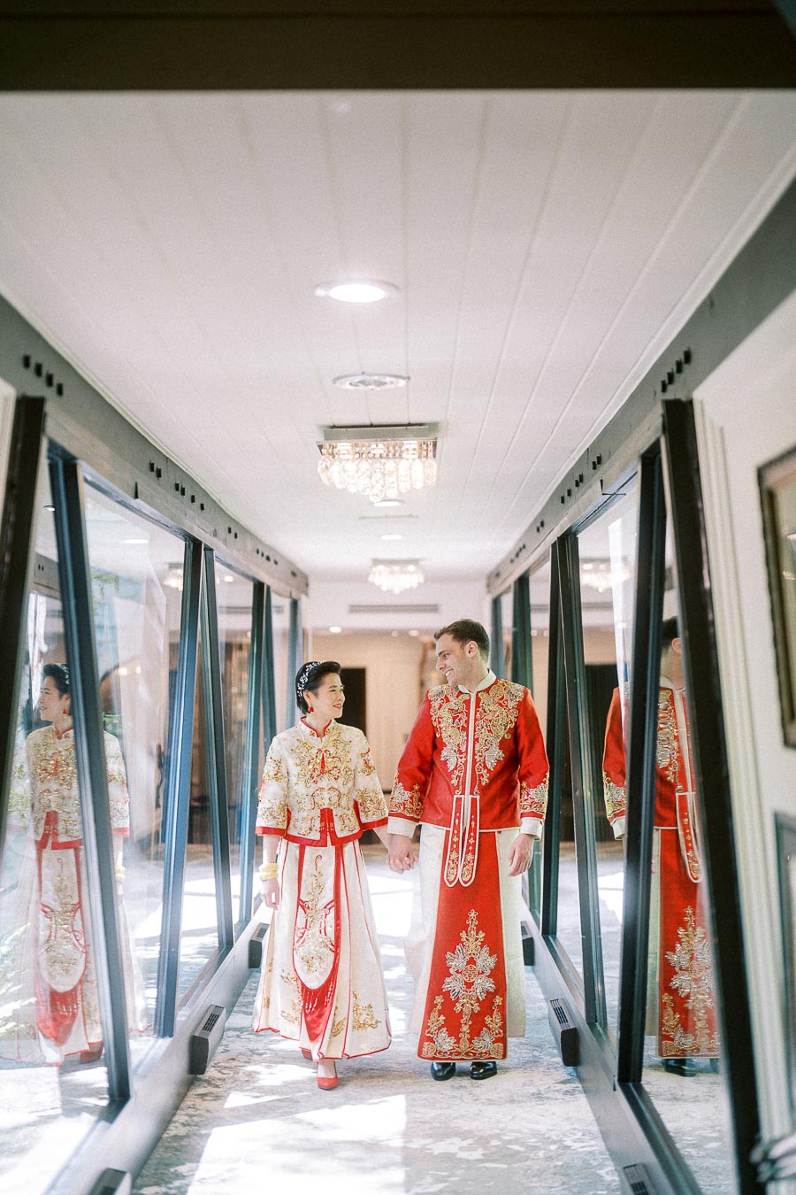 Miffy and Rawle hold hands in red and gold Chinese wedding outfits while walking through a glass corridor.