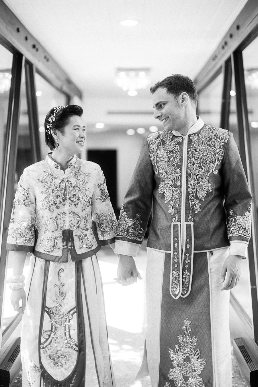 Miffy and Rawle hold hands in ornate traditional Chinese wedding outfits while smiling in a bright hallway.