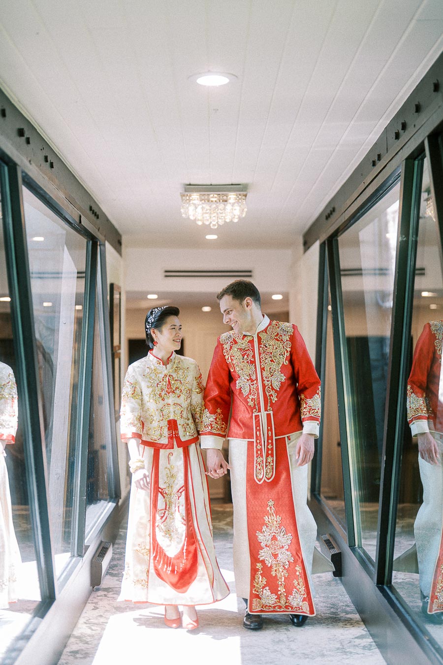 Miffy and Rawle holding hands in red and gold Chinese wedding outfits in a bright glass hallway.