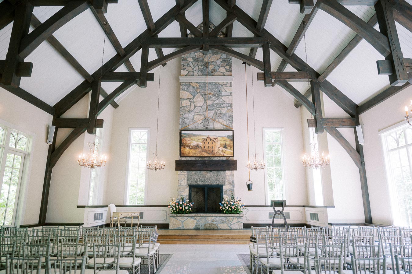Empty chapel ceremony room with timber beams, chandeliers, rows of chairs, stone fireplace, and floral arrangements