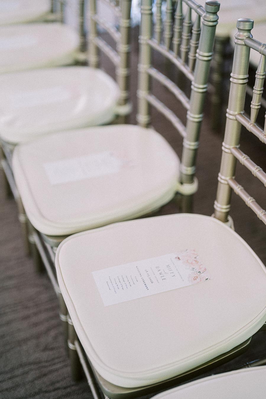 Program cards rest on white chiavari ceremony chairs with silver frames at Miffy and Rawle's wedding.