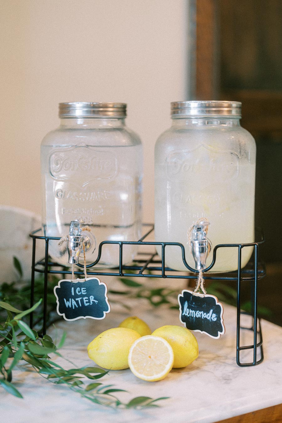 Glass drink dispensers labeled ice water and lemonade with lemons and greenery on a marble table.