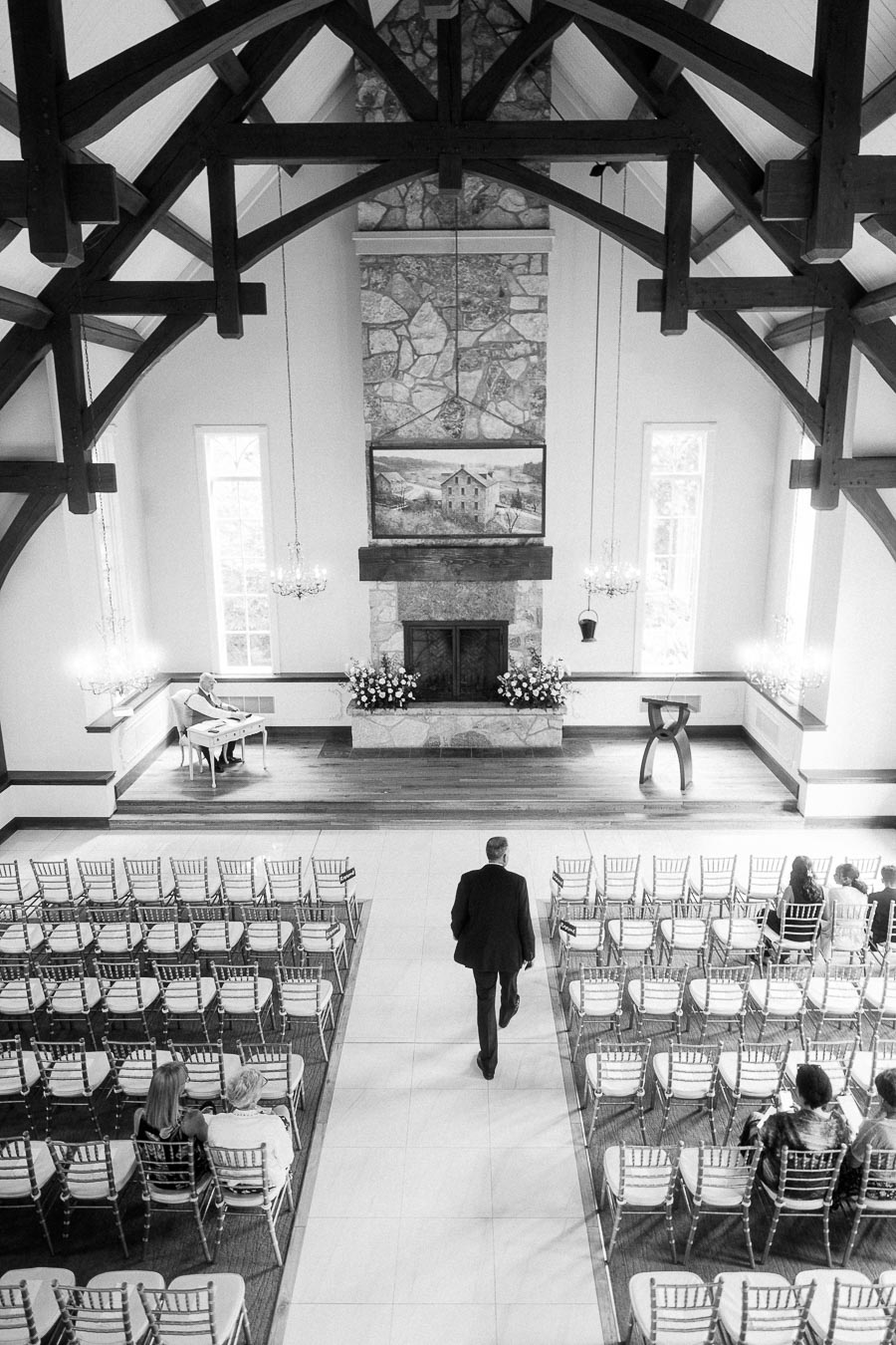 Groom walks down the aisle inside Ancaster Mill chapel with timber beams, stone fireplace, piano, and seated guests.