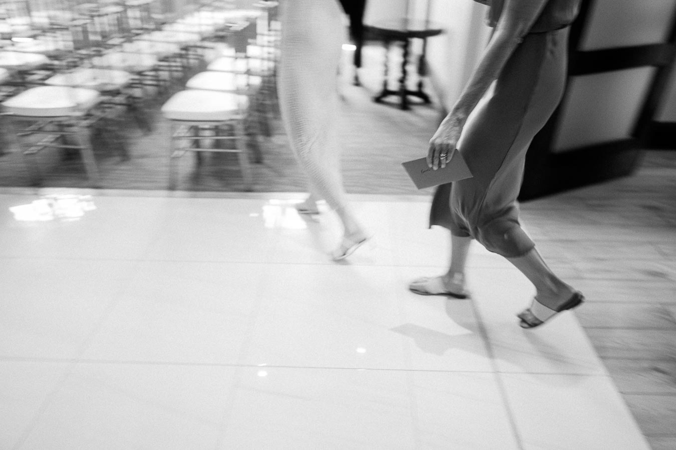 Black and white motion blur of two people walking across a glossy floor near a banquet room.