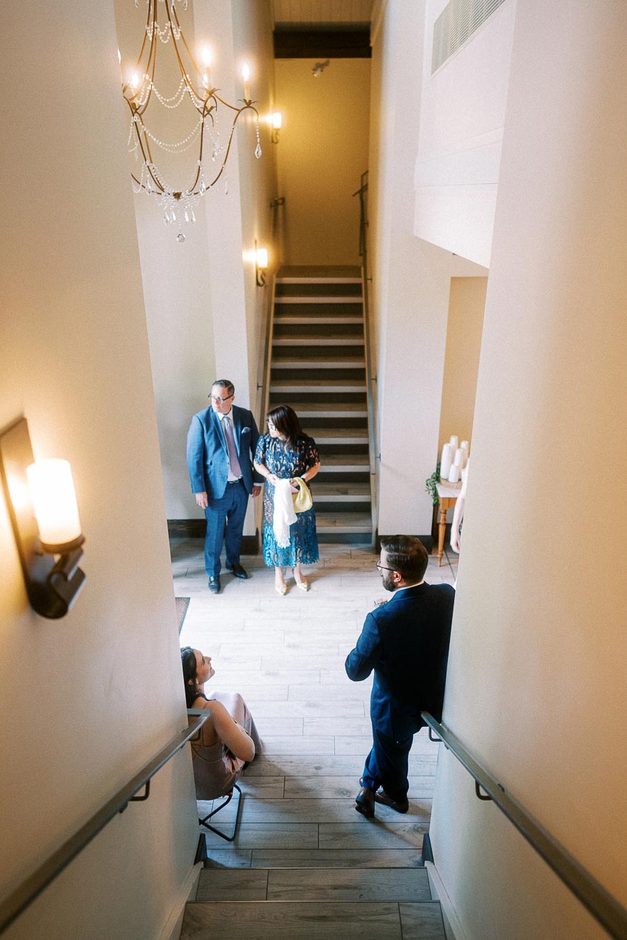 Wedding guests talk in a tall stairwell with chandelier and wall sconces at Ancaster Mill.