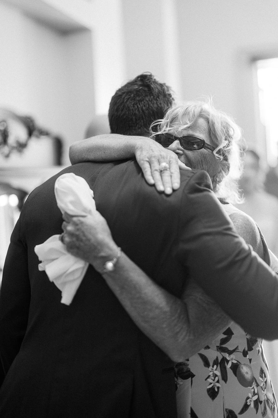 Black and white close-up of an older woman in sunglasses hugging a man in a suit indoors.