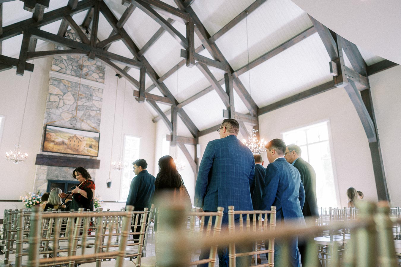 Wedding guests stand among chiavari chairs inside a bright chapel with timber beams and a stone fireplace.