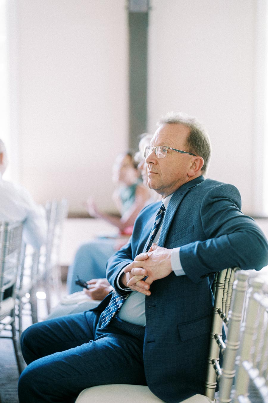 Man in a blue suit and glasses sits on a white chair among wedding guests in a bright ceremony room.