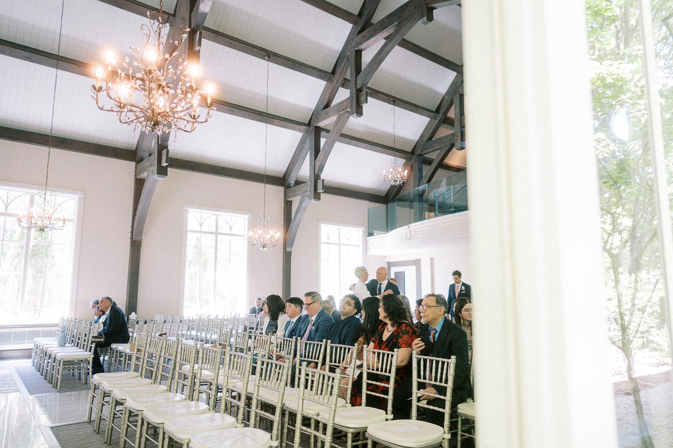 Guests sit in white chairs inside a bright chapel with chandeliers, dark beams, and tall windows.