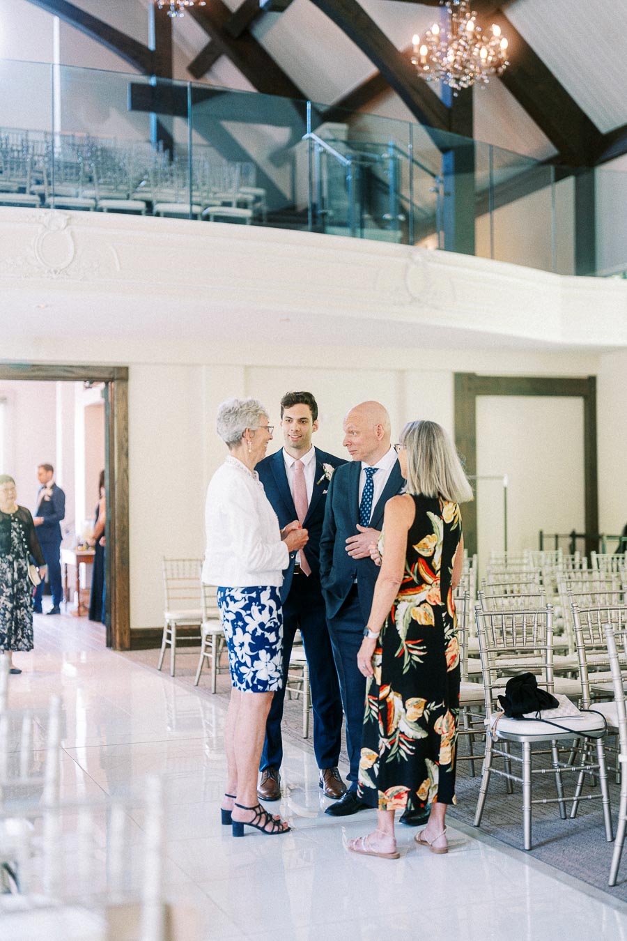 Wedding guests talk beside rows of ceremony chairs in a bright hall with chandeliers and a glass balcony.