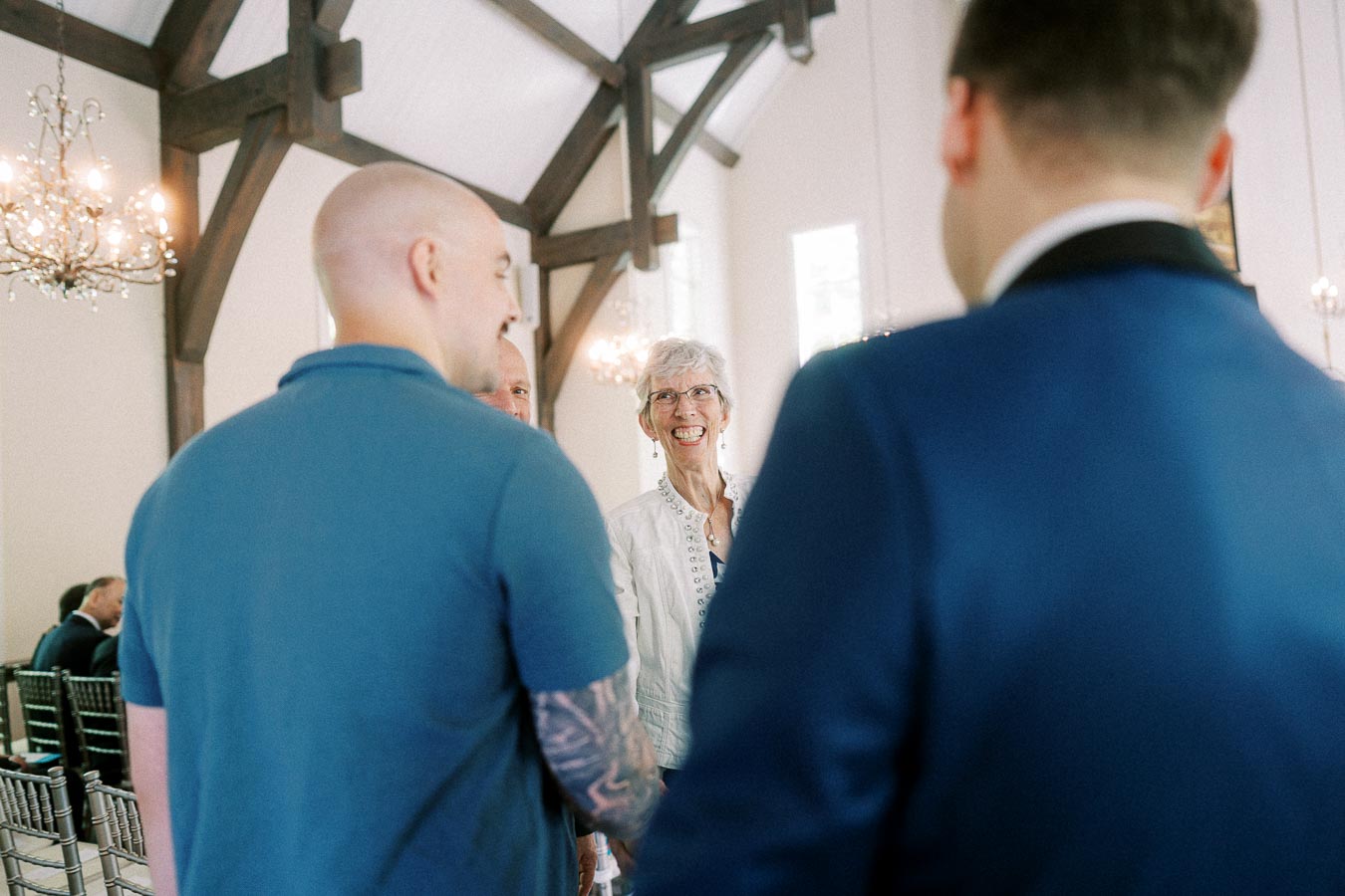 Guests chat and smile inside Ancaster Mill chapel with chandeliers, timber beams, and ceremony chairs.